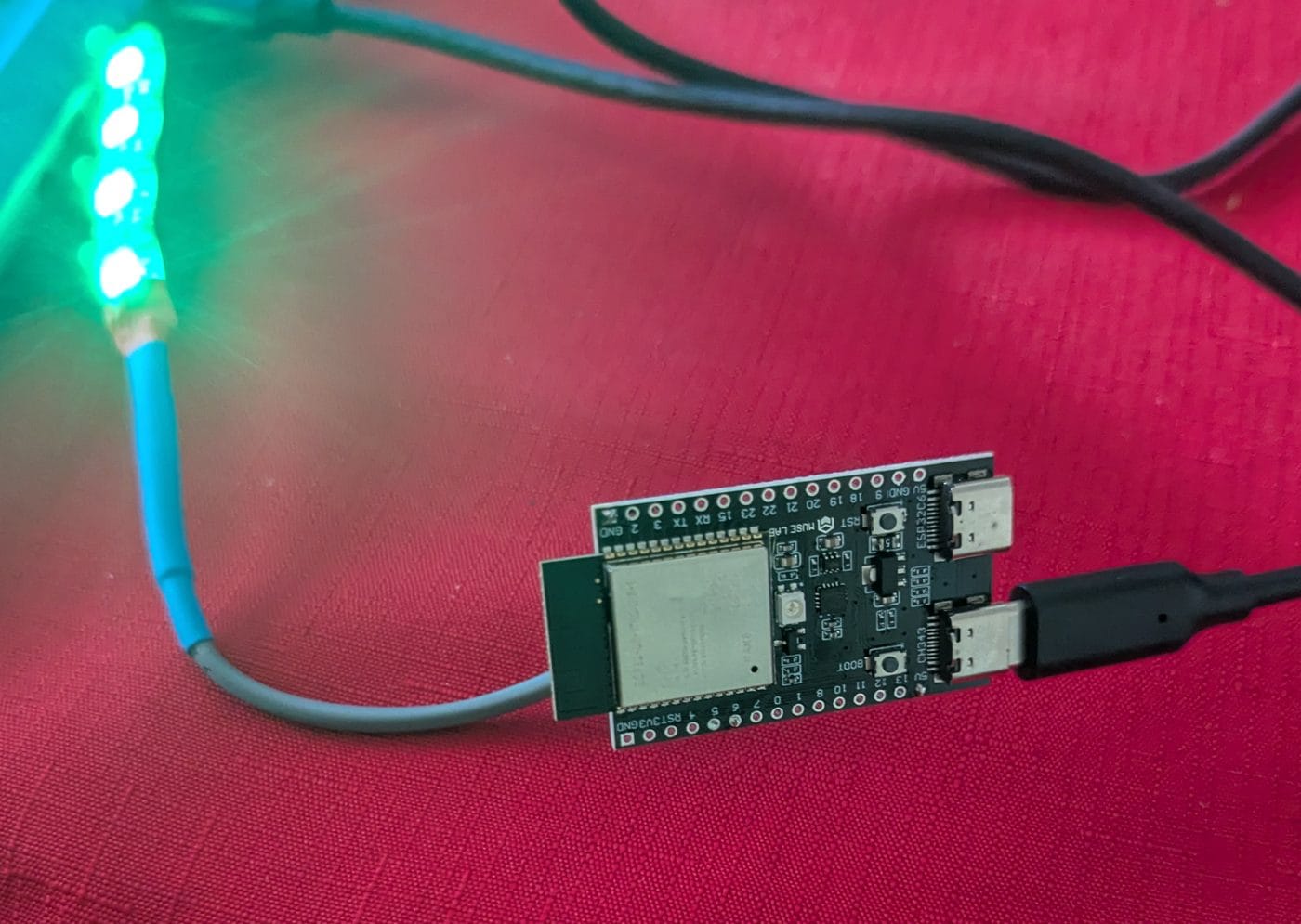 ESP32-C6 board on a red cloth with its WS2812B strip glowing green, USB-connected to a laptop