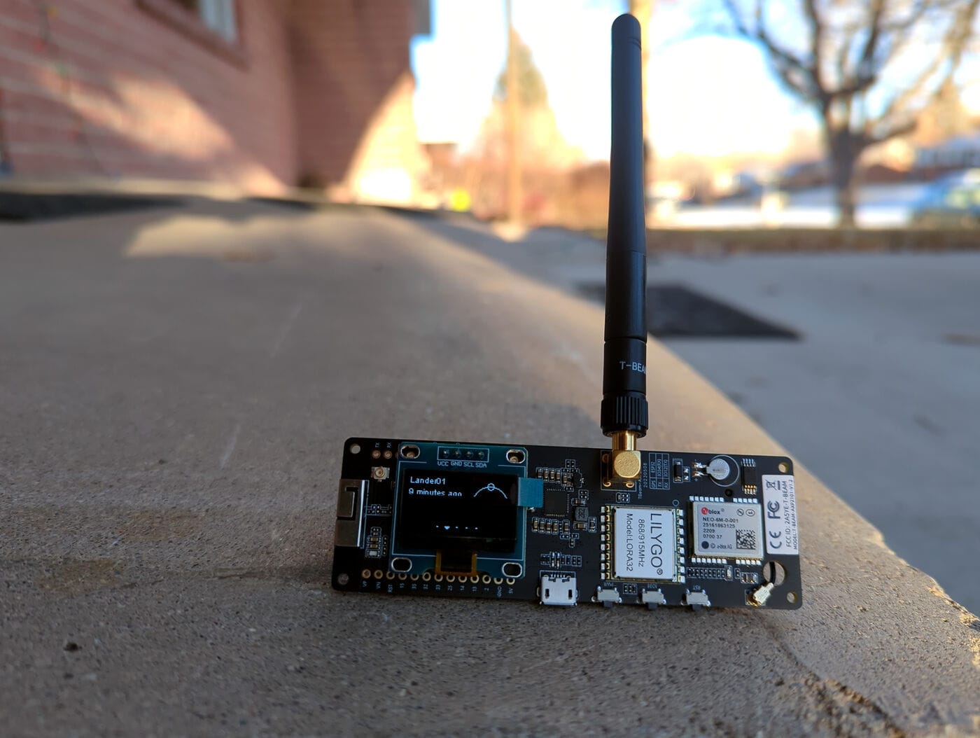A LilyGO T-Beam board standing upright on a concrete ledge, showing its LILYGO LORA32 868/915MHz radio module, GPS module, OLED display, and antenna